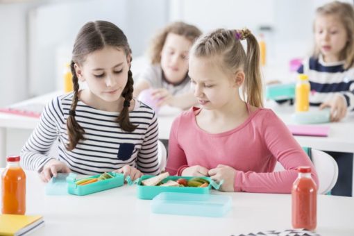 Two young girls during snack time in a school looking into each other's lunch boxes with healthy vegetables and bread. Bottles of fruit juices on the desk. Other kids in blurred background